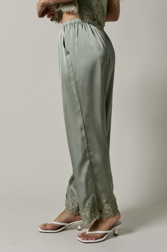 A person wearing wide-leg sage green satin pants with lace trim at the hem, paired with white heeled sandals, shown against a plain light gray studio background.