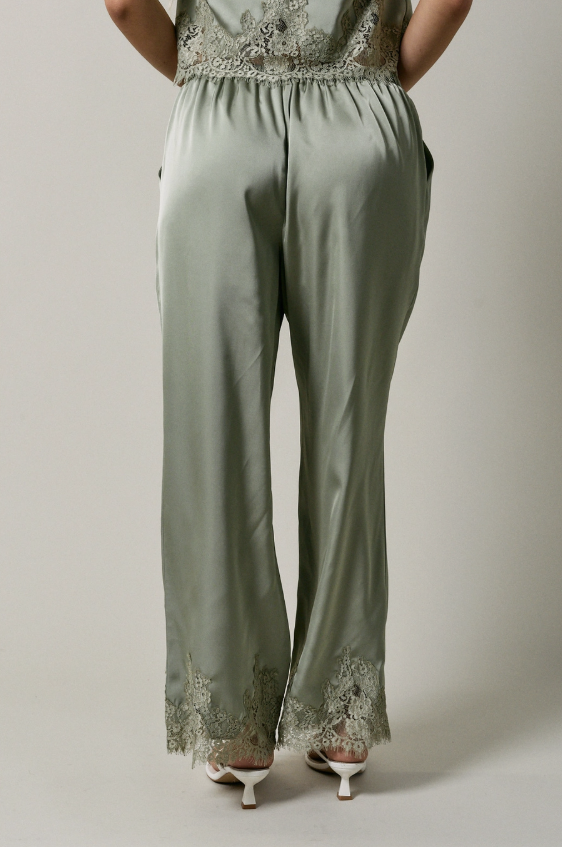 A person shown from the back wearing wide-leg sage green satin pants with floral lace trim at the waist and hems, paired with white heeled sandals against a plain light gray studio background.
