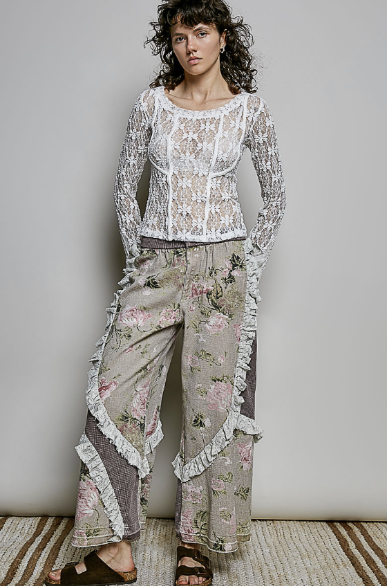 A model wearing a bohemian-style two-piece outfit featuring a white lace long-sleeve crop top and wide-leg beige pants with pink floral prints and ruffle trim, standing against a neutral gray wall.
