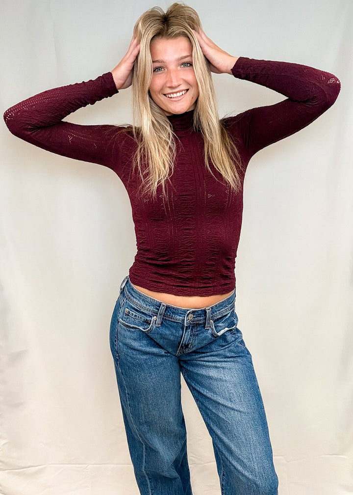 Woman wearing burgundy mock neck top with blue jeans on white background