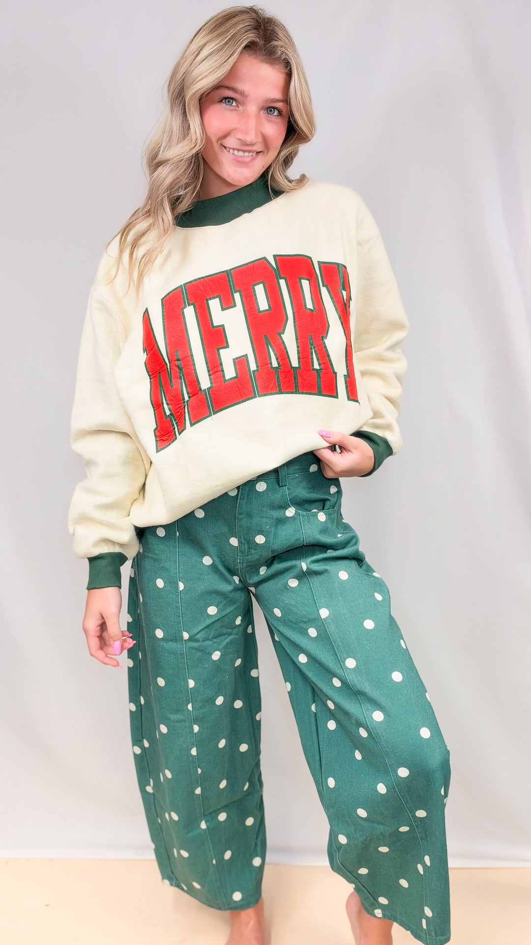 Woman wearing green polka dot barrel pants with cream mock neck sweatshirt with 'MERRY' print on front on plain background