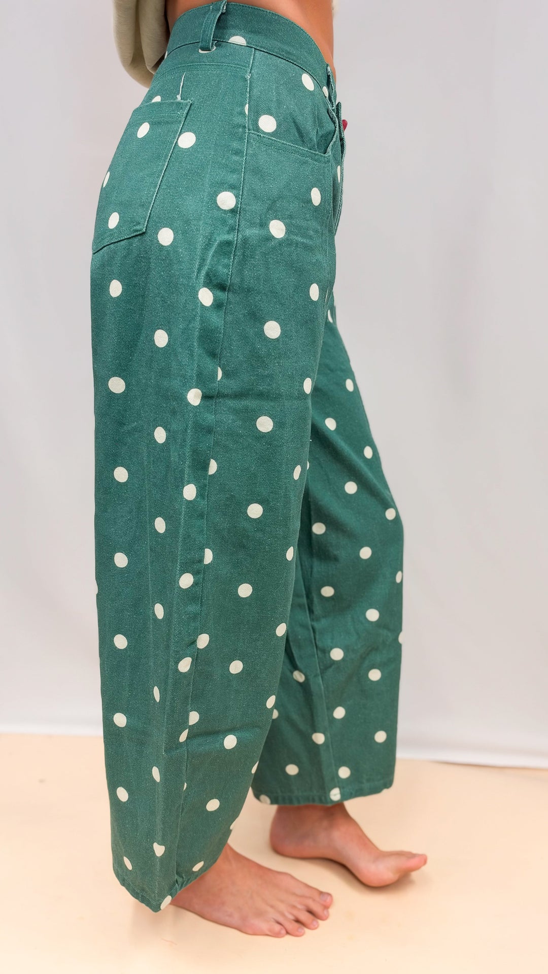 Woman shown from side view wearing green polka dot barrel jeans on plain background