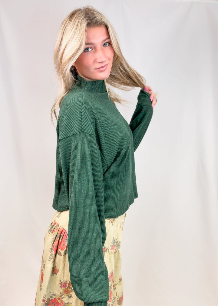 Woman wearing green mock neck sweater and floral pants on plain background