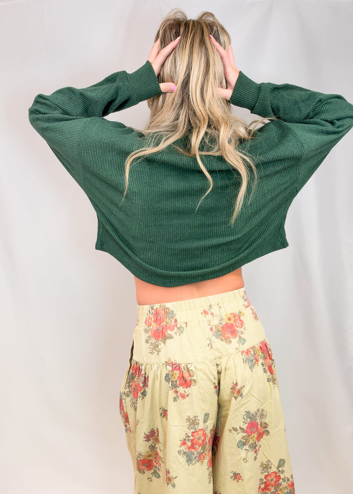 Woman shown from back view wearing green sweater and floral pants on plain background