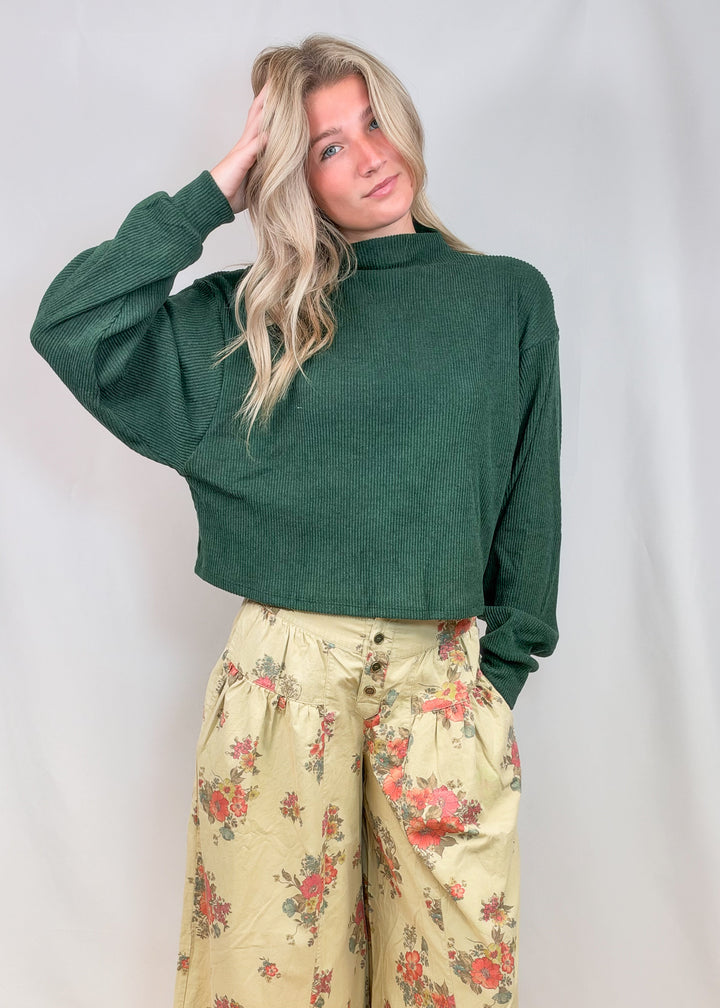 Woman wearing green mock neck sweater with floral pants on plain background