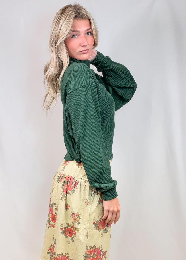 Woman shown from side view wearing green sweater and floral pants on plain background