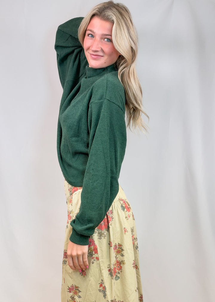 Woman wearing green mock neck sweater and floral pants on plain background