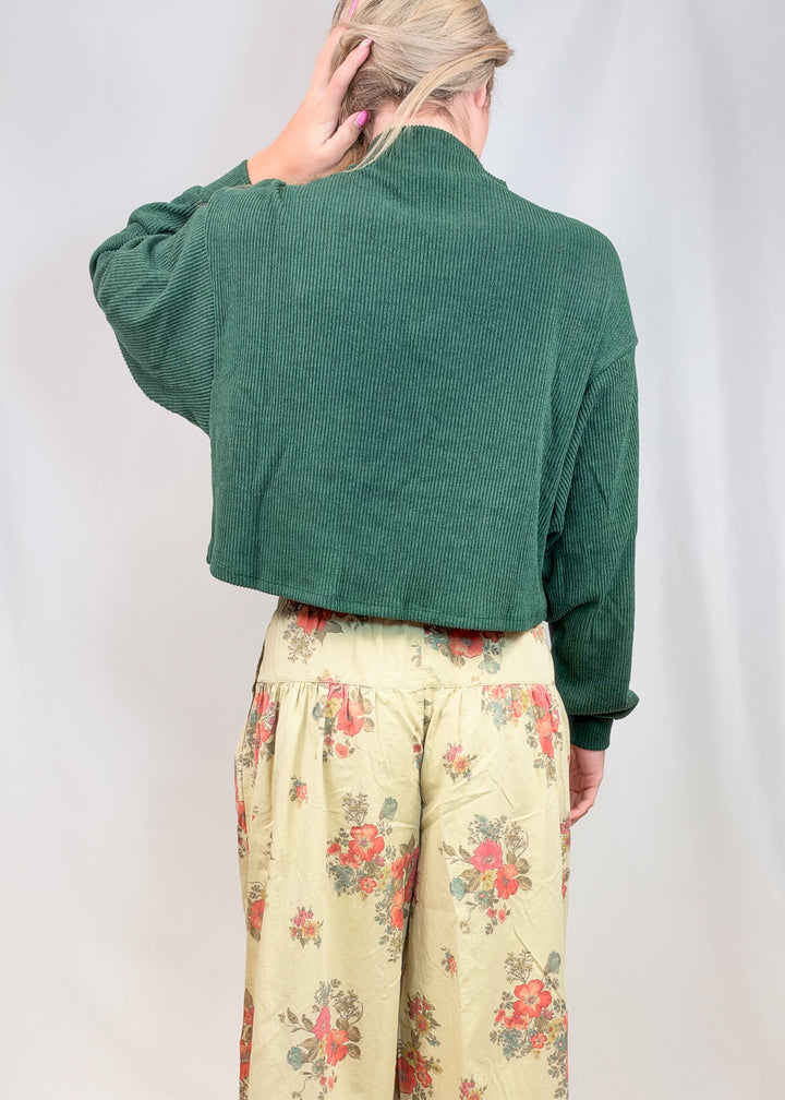 Woman shown from back wearing green sweater and floral pants on plain background