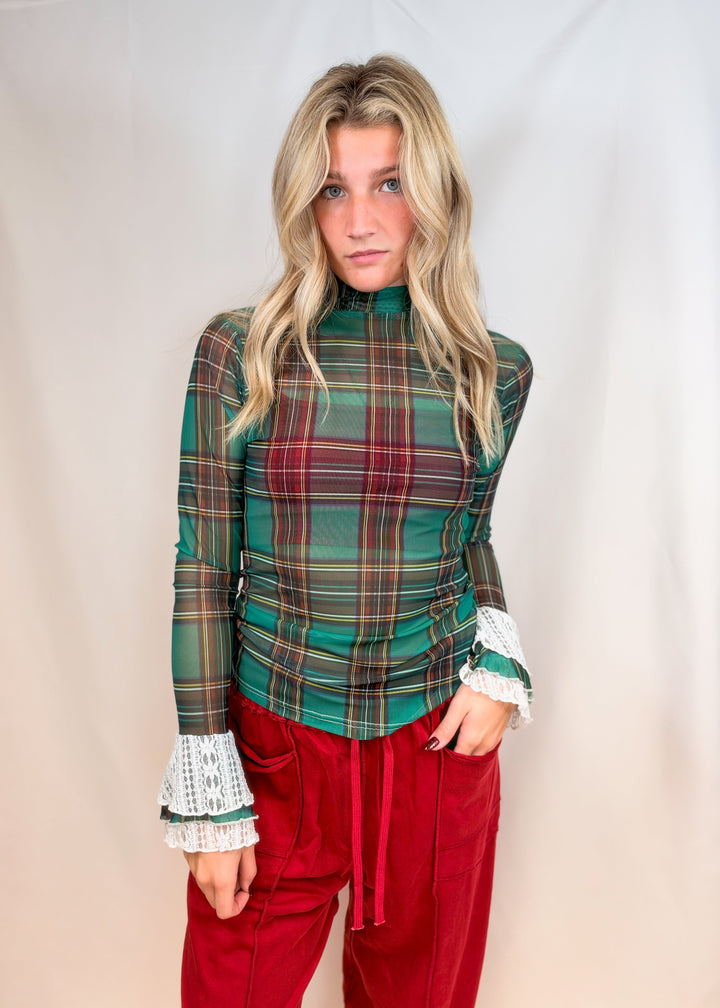 Woman wearing green and red tartan sheer top with lace cuff sleeves and red pants on plain background