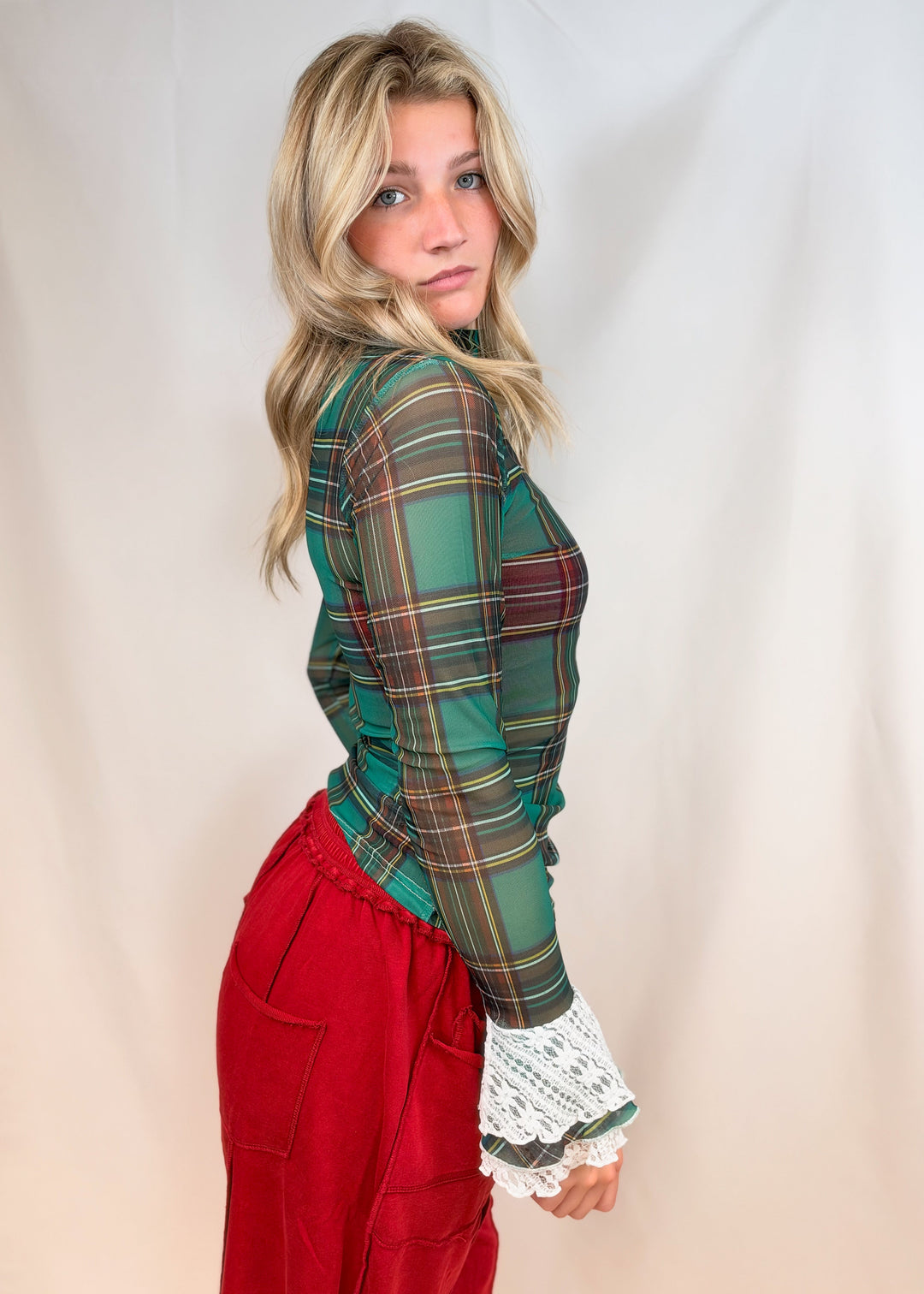 Woman shown from side view wearing green and red tartan sheer top with lace cuff sleeves and red pants on plain background