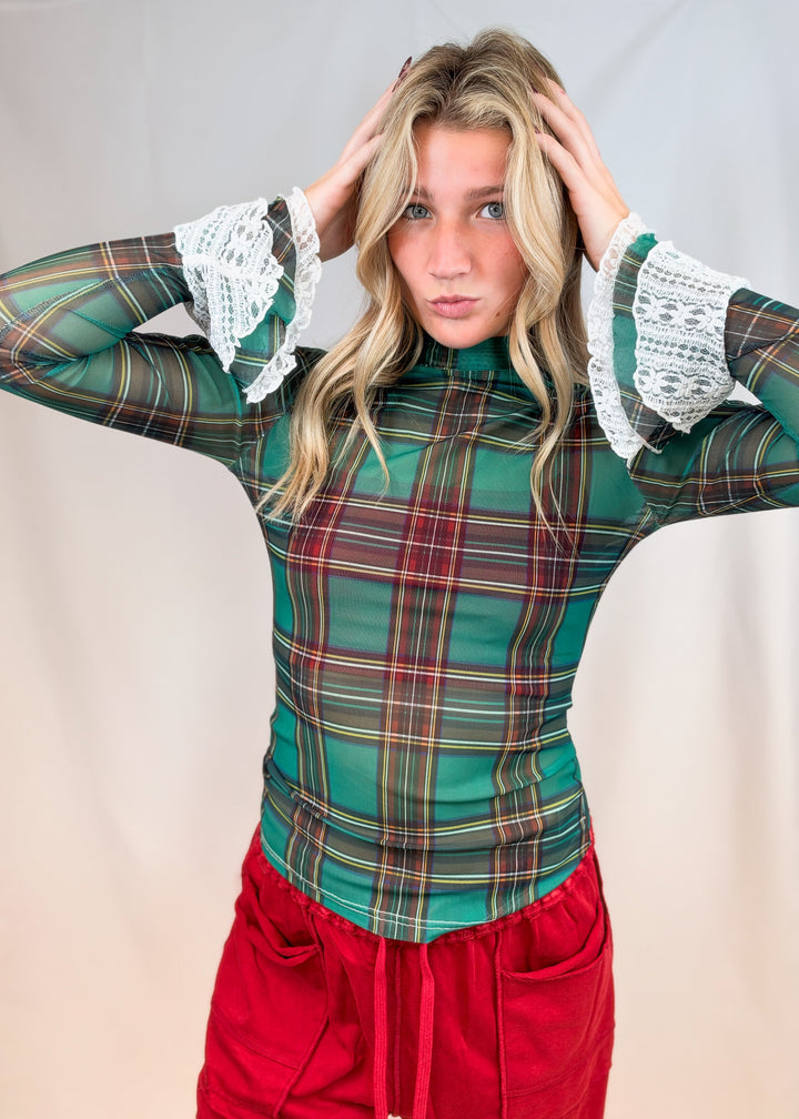 Woman wearing green and red tartan sheer top with lace cuff sleeves and red pants on plain background