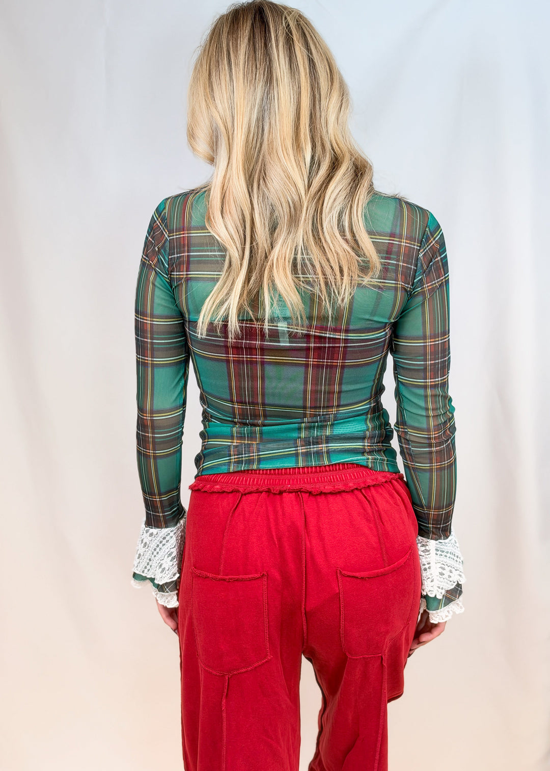 Woman shown from back view wearing green and red tartan sheer top with lace cuff sleeves and red pants on plain background