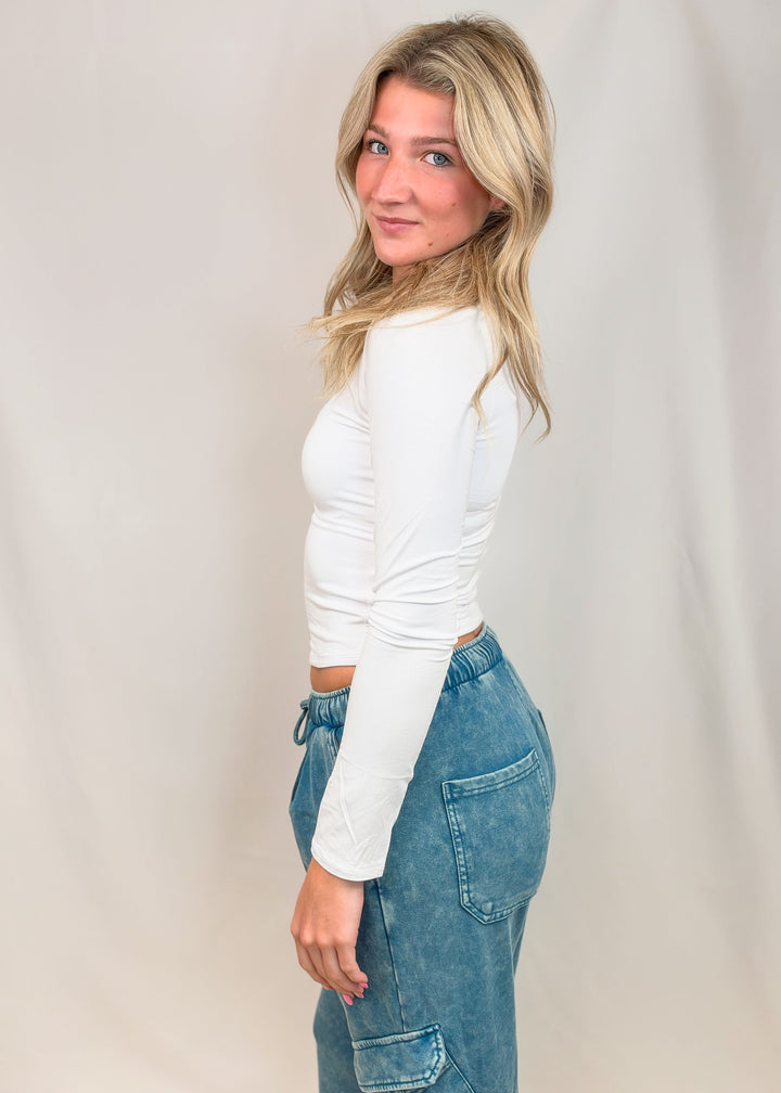 Woman shown from side wearing white long sleeve top on plain background