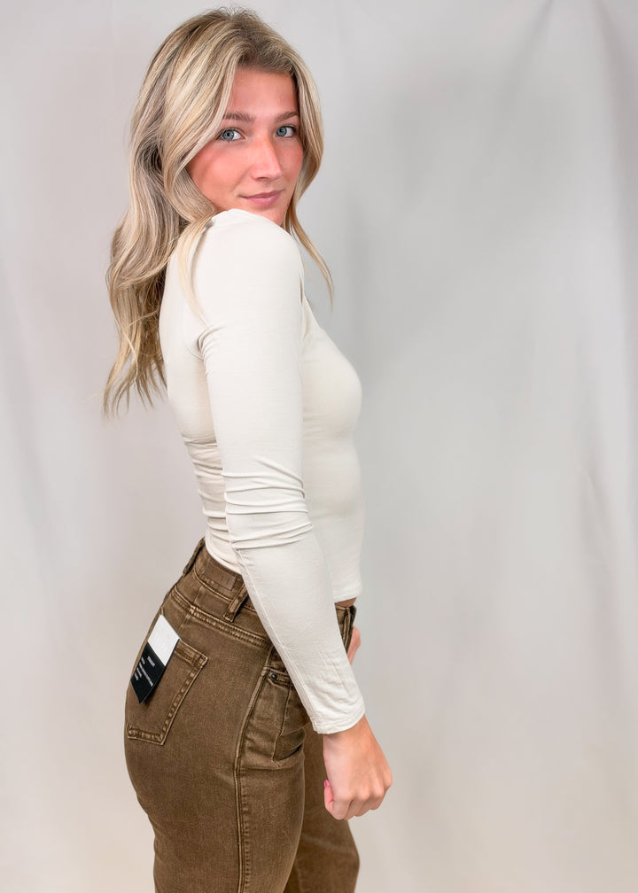 Woman shown from side view wearing cream long sleeve top on plain background