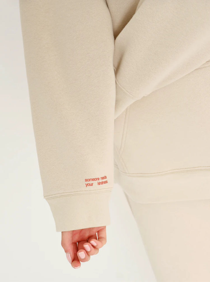 Beige hoodie with red text on sleeve against a white background