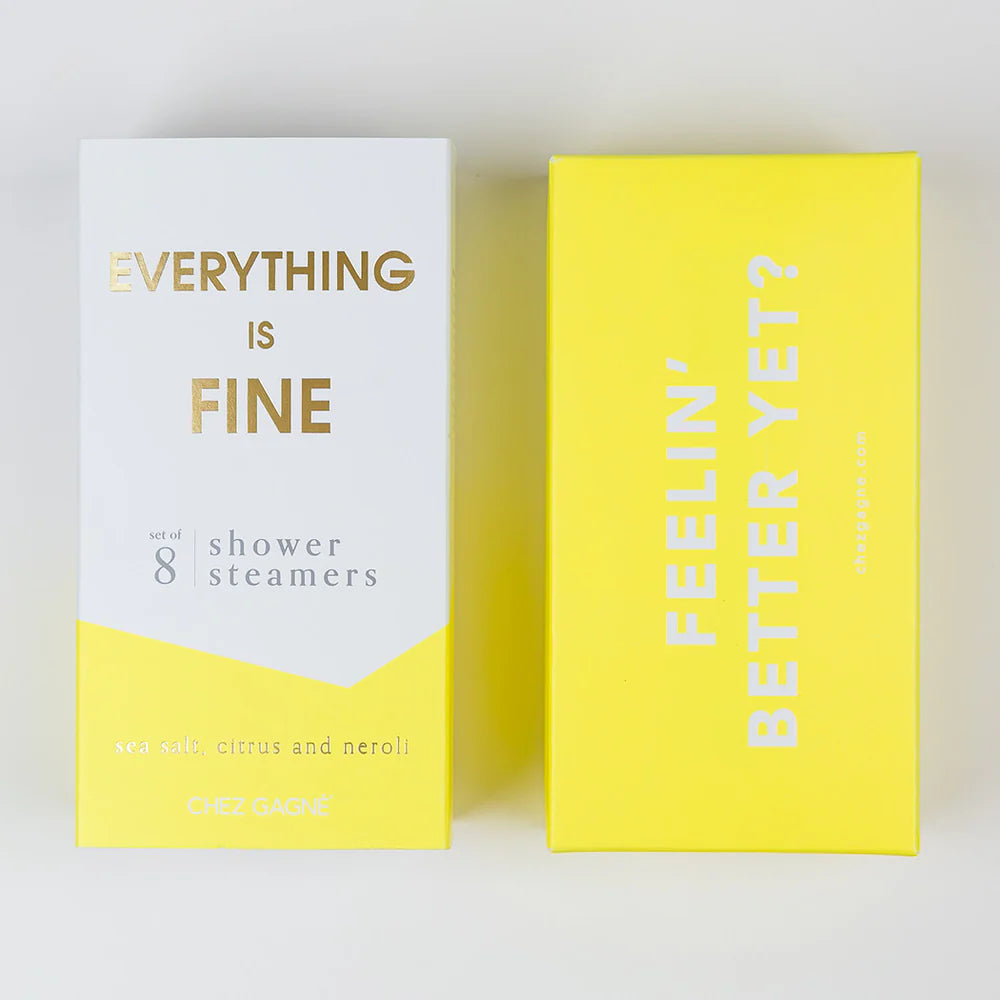 Two product boxes for shower steamers on a light neutral background. The left box is white with a yellow lower section and gold text reading &