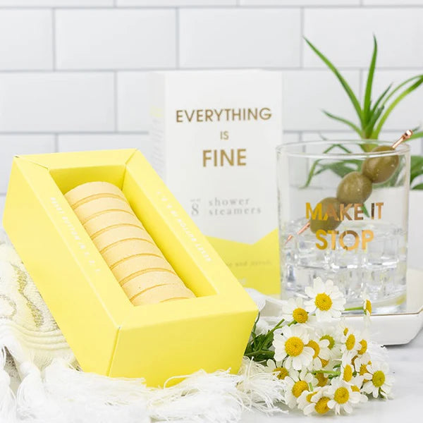 A yellow box of shower steamers next to its white and gold packaging box, styled with a glass of water, chamomile flowers, and a small plant on a white tiled background.