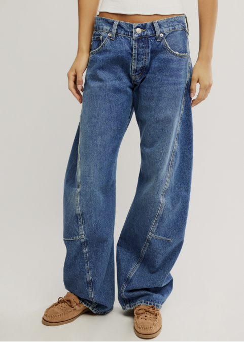 A person wearing wide-leg blue denim jeans with a white cropped top and brown lace-up shoes, standing against a light gray studio background.