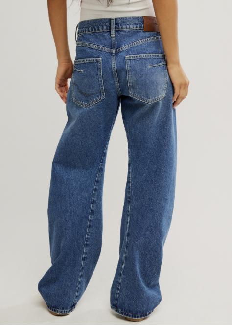 A person shown from the back wearing wide-leg blue denim jeans with a white ribbed top, photographed against a plain light gray studio background.