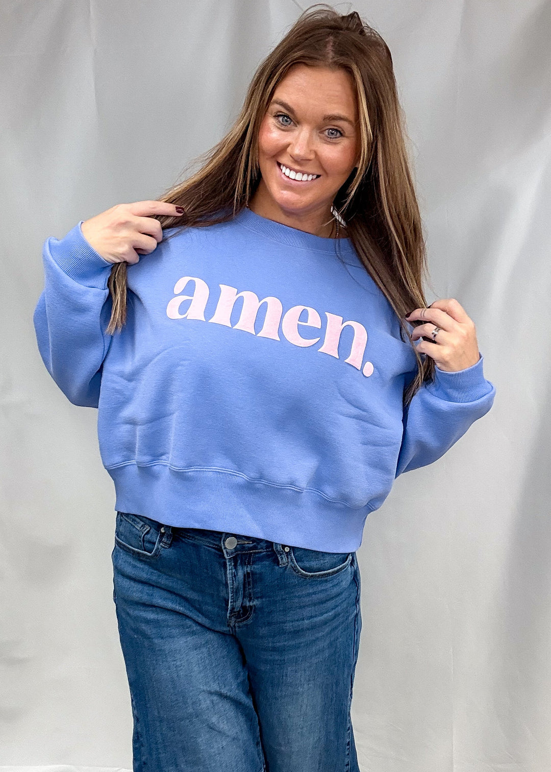 A woman modeling a light blue crewneck sweatshirt with the word 'amen.' printed in white across the chest, paired with blue jeans, standing against a white fabric backdrop.