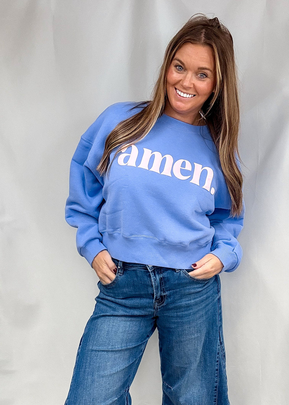 A woman wearing a light blue crewneck sweatshirt with the word 'amen.' printed in white across the chest, paired with wide-leg blue jeans, standing against a white fabric backdrop.