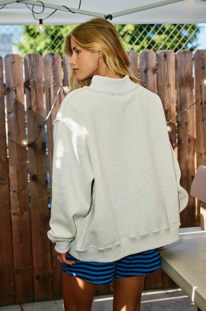 Woman wearing a light gray sweatshirt and blue shorts standing outdoors with a wooden fence in the background.