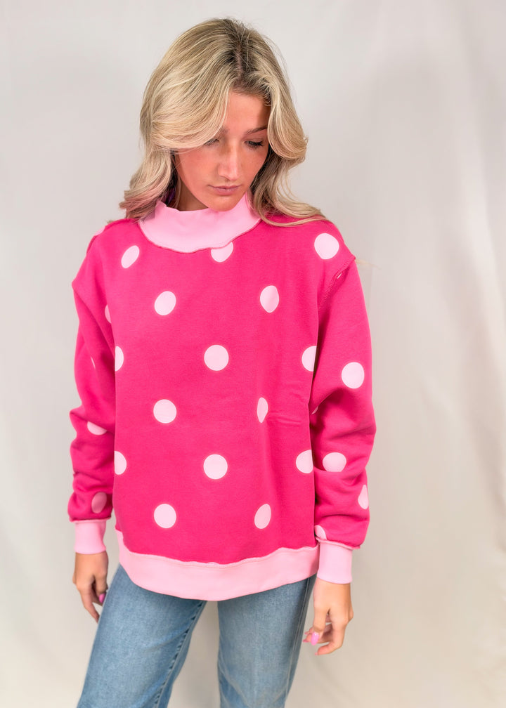 Woman wearing pink mock neck sweatshirt with light pink polka dots with jeans on plain background