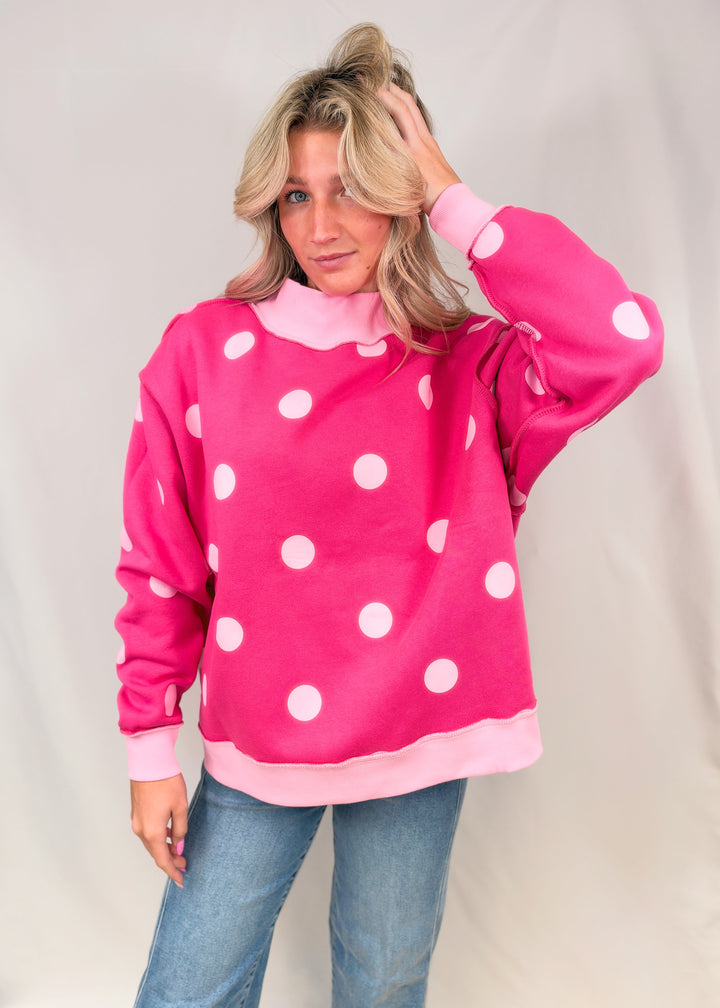 Woman wearing pink mock neck sweatshirt with light pink polka dots and denim jeans on plain background