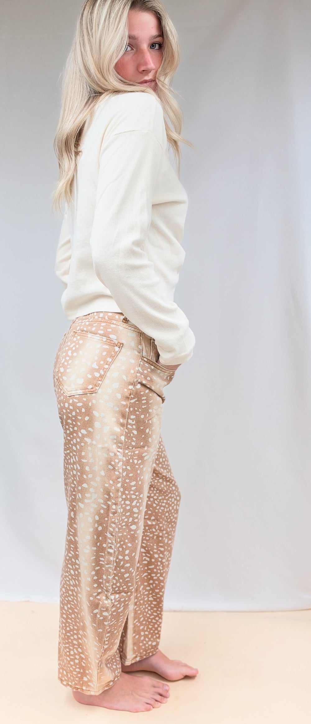 Woman shown from side view wearing beige color top with fawn print denim barrel jeans on plain background