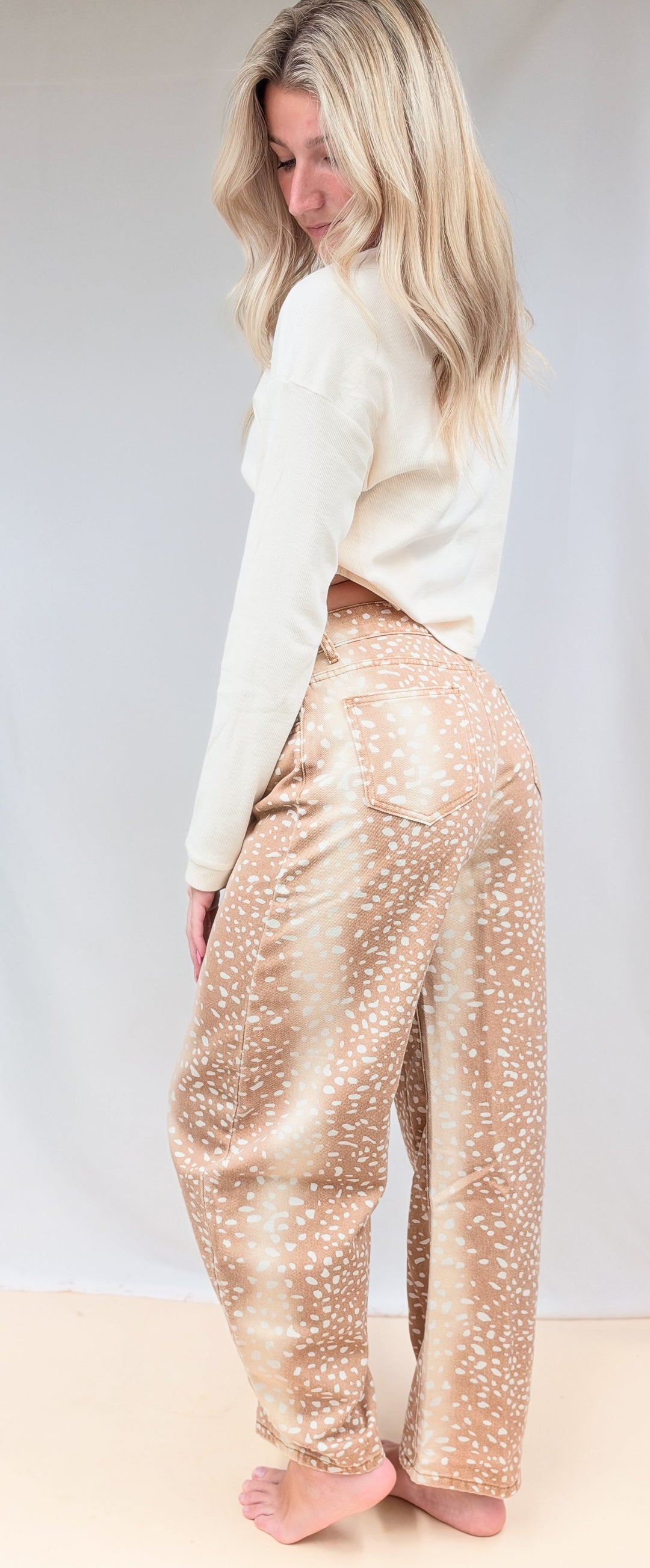 Woman shown from side view wearing beige color top with fawn print denim barrel jeans on plain background