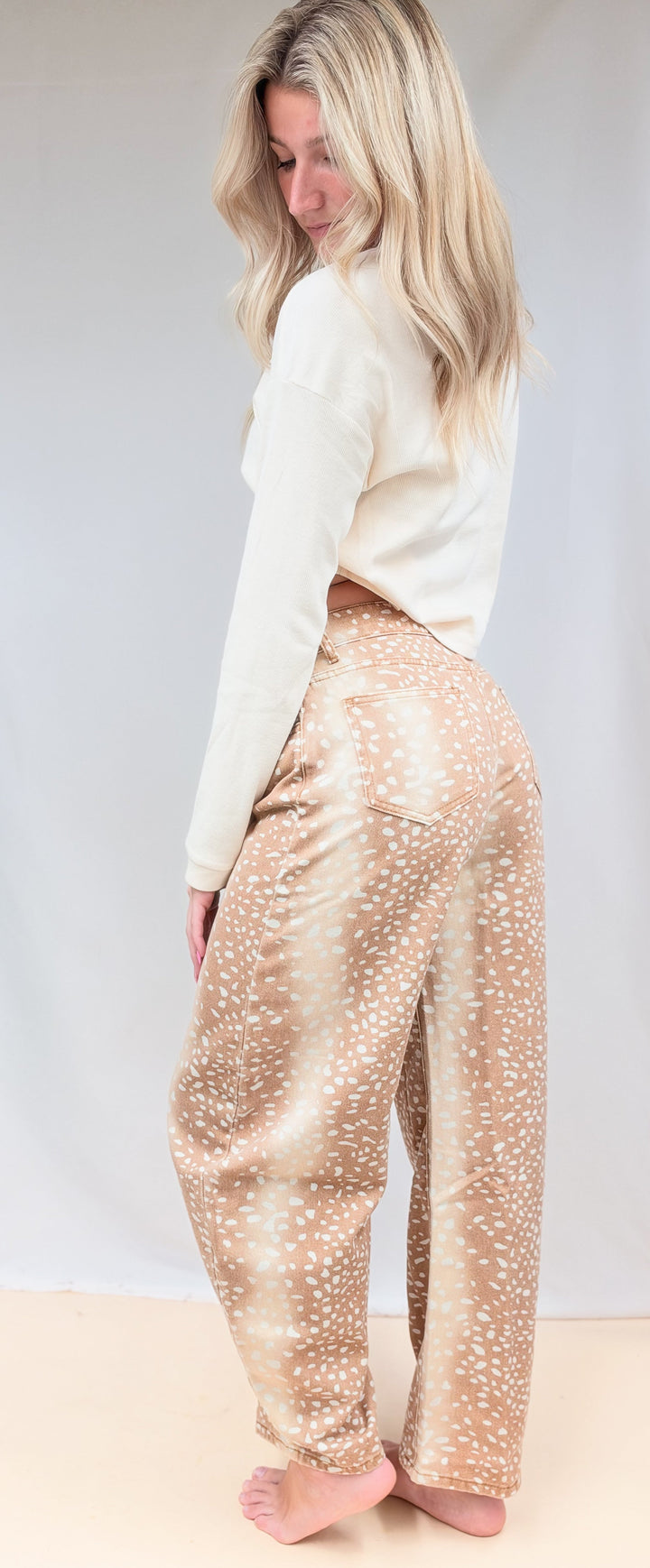 Woman shown from side view wearing beige color top with fawn print denim barrel jeans on plain background