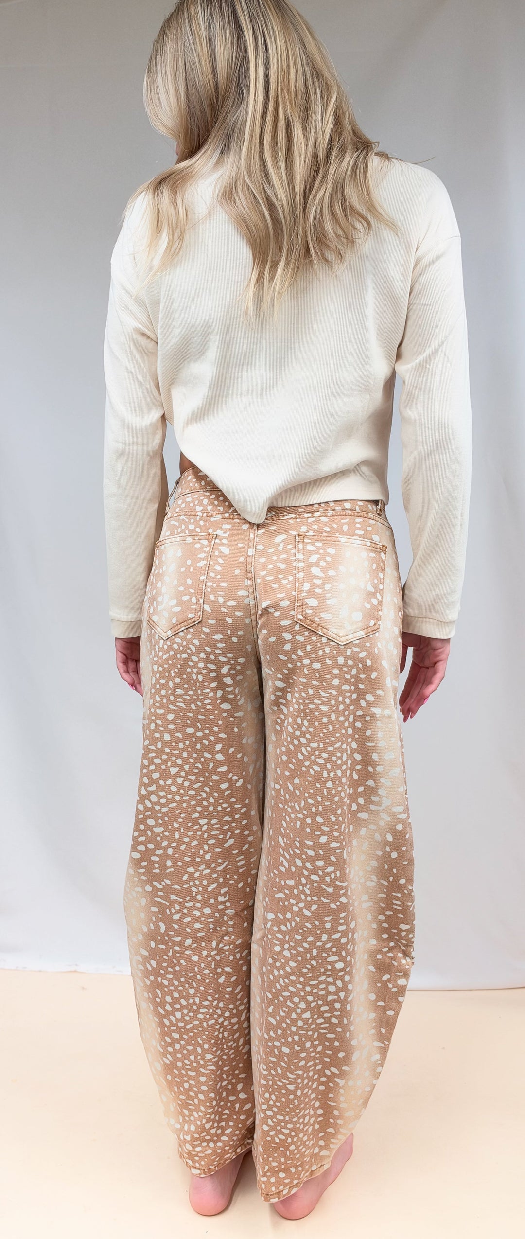 Woman shown from back view wearing beige color top with fawn print denim barrel jeans on plain background
