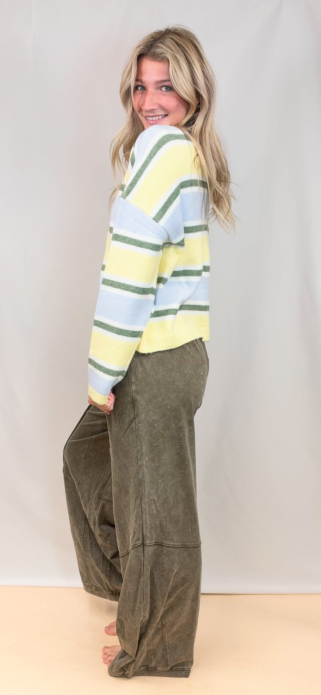 Woman shown from side view wearing yellow, green and blue stripe cardigan with green pants on plain background