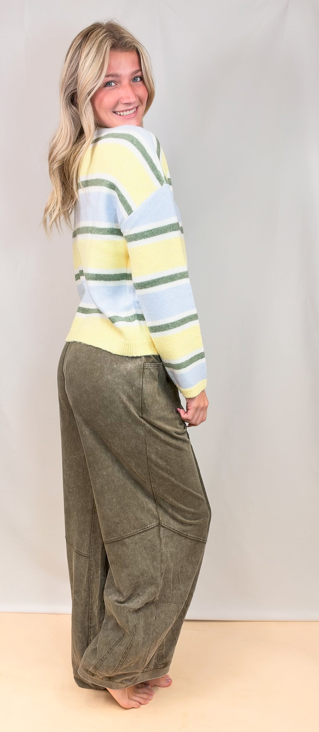 Woman shown from side view wearing yellow, green and blue stripe cardigan with green pants on plain background
