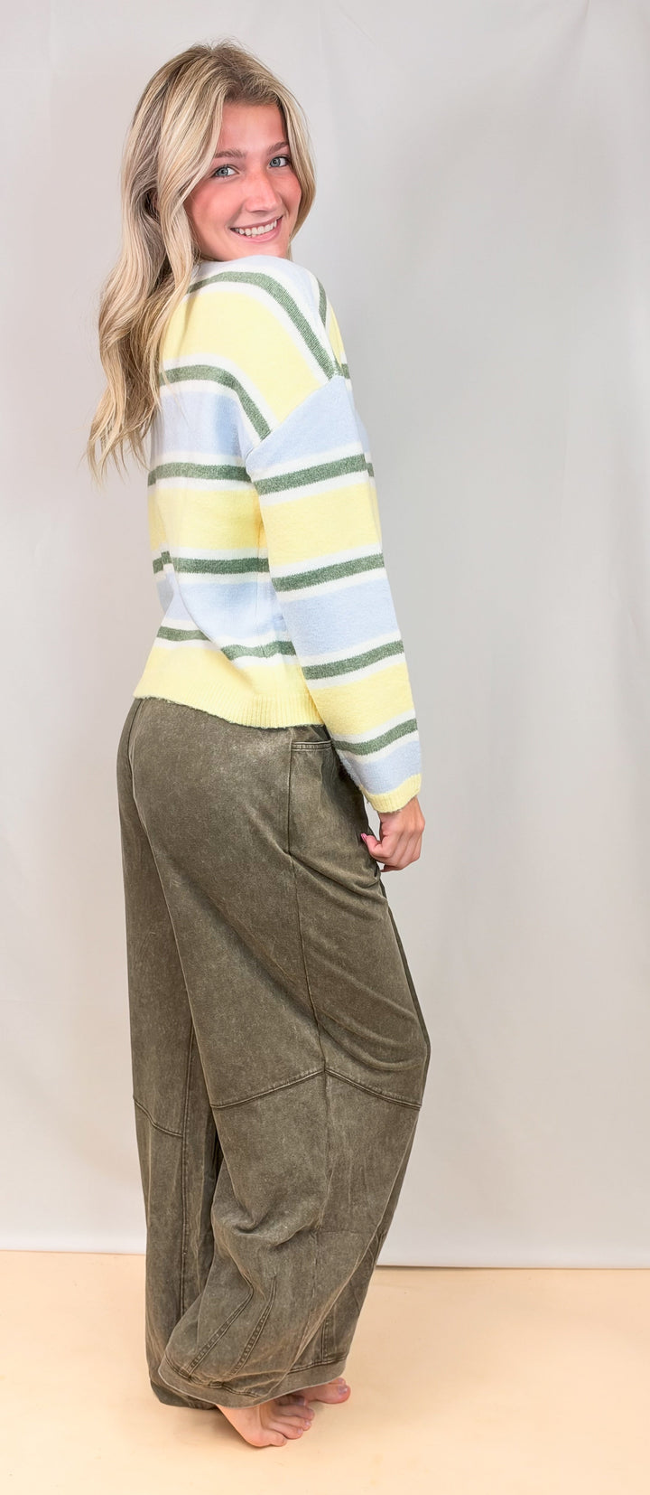 Woman shown from side view wearing yellow, green and blue stripe cardigan with green pants on plain background