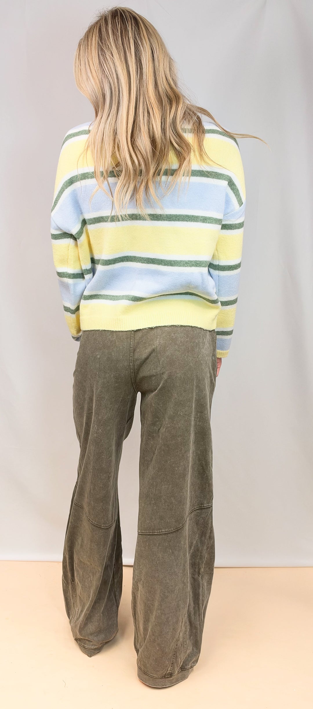 Woman shown from back view wearing yellow, green and blue stripe cardigan with green pants on plain background