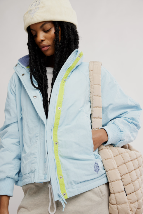 Person wearing a light blue jacket with a hood, holding a beige quilted bag against a plain background