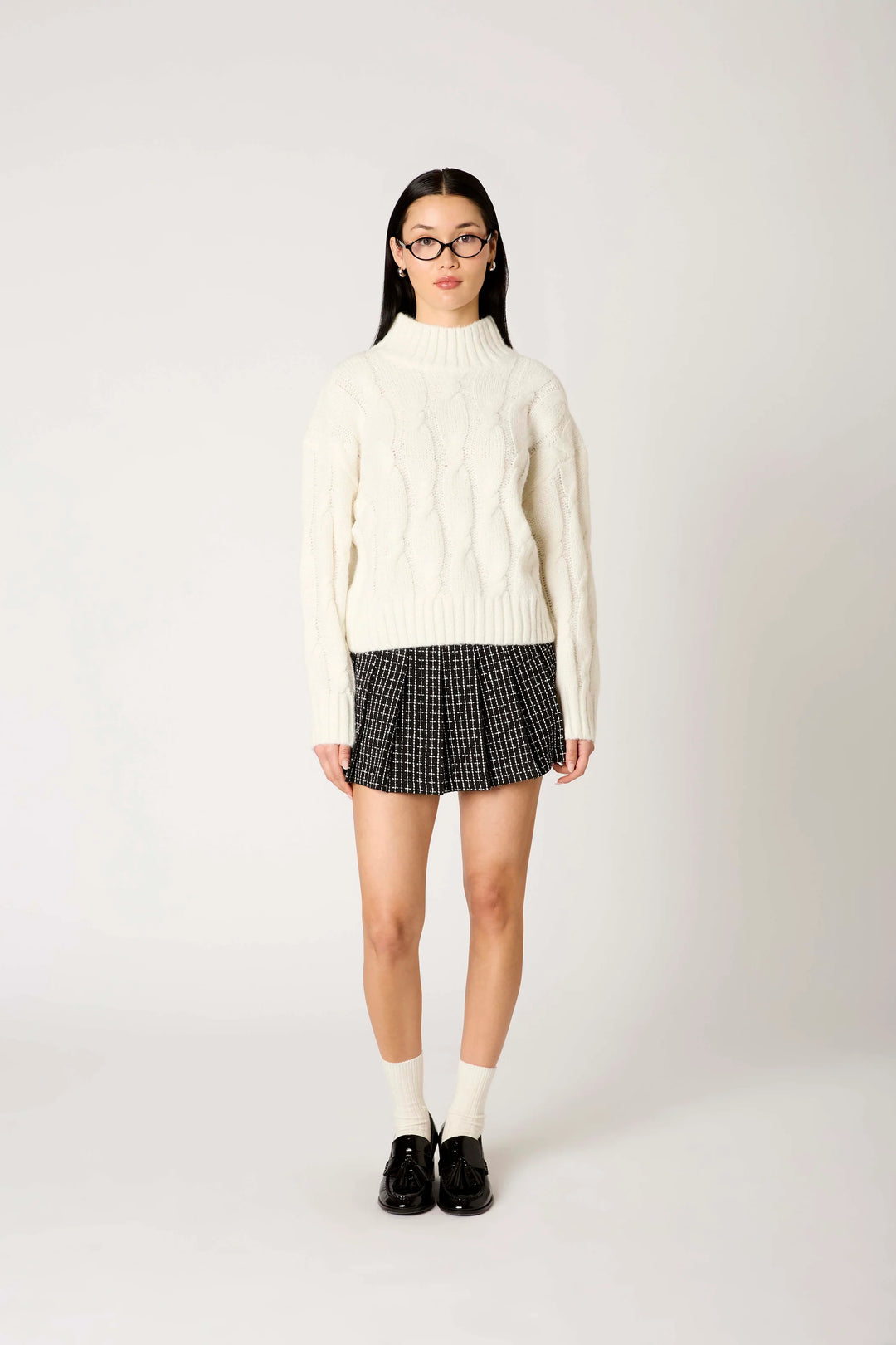 A model stands against a light gray studio background wearing a cream cable-knit mock-neck sweater, a black and white checkered pleated mini skirt, white socks, and black loafers.
