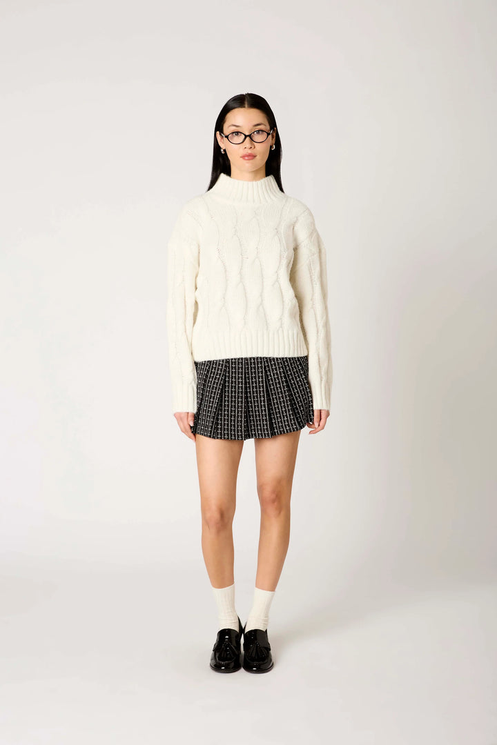 A model stands against a light gray studio background wearing a cream cable-knit mock-neck sweater, a black and white checkered pleated mini skirt, white socks, and black loafers.