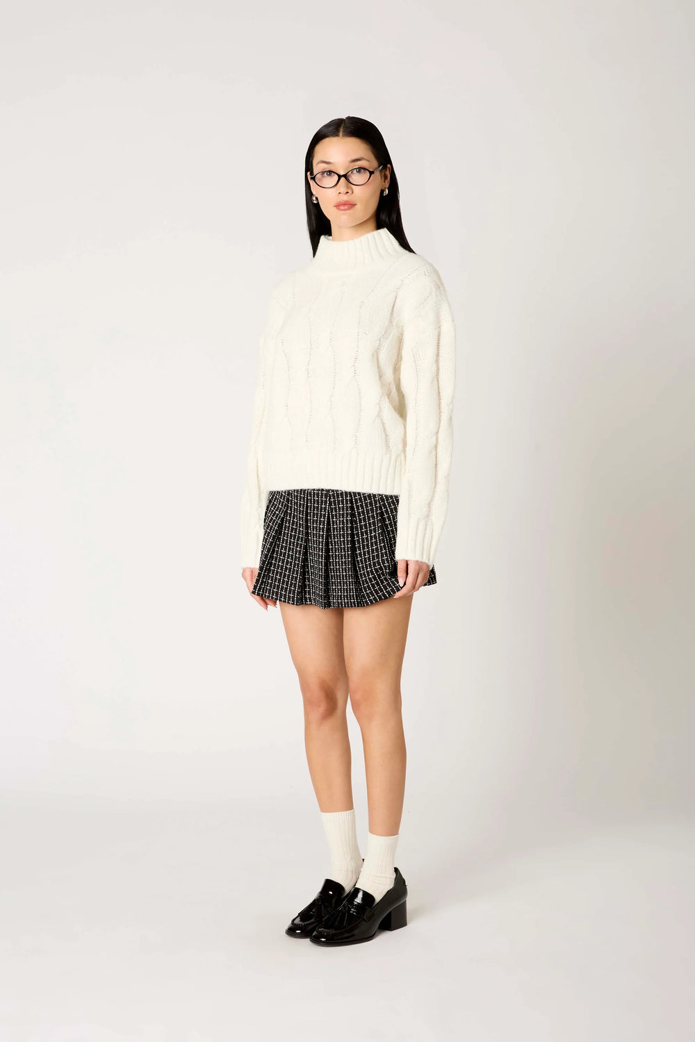 A model wearing a cream cable-knit mock-neck sweater, a black and white checkered pleated mini skirt, white socks, and black loafers, standing against a light gray studio background.