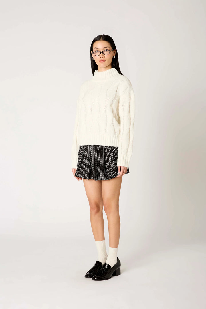 A model wearing a cream cable-knit mock-neck sweater, a black and white checkered pleated mini skirt, white socks, and black loafers, standing against a light gray studio background.