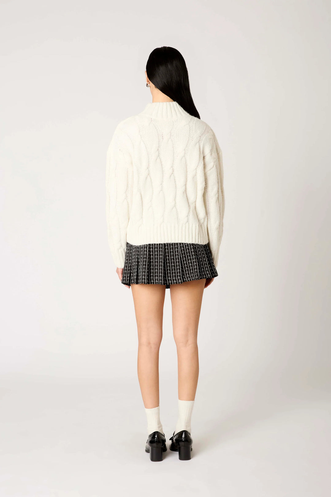 Person wearing a white cable knit sweater and plaid skirt on a white background