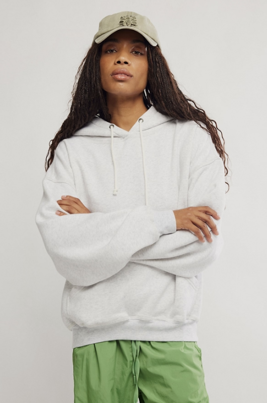 A model wearing an oversized light gray heather pullover hoodie with white drawstrings, a beige baseball cap, and green pants, standing against a plain light gray studio background.