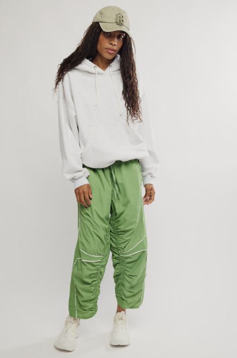 A model wearing a white pullover hoodie, sage green ruched cargo pants, a beige baseball cap, and white sneakers, standing against a light gray studio background.