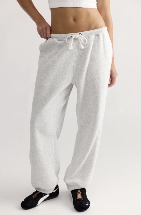 A person wearing light heather gray sweatpants with a white drawstring, paired with black sneakers, shown from the waist down against a plain light gray studio background.