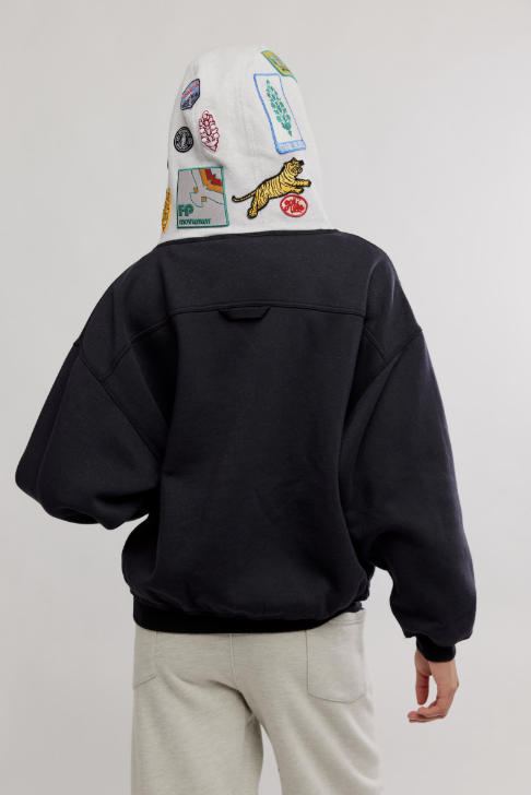 Person wearing a black jacket and light-colored pants with a white beanie featuring colorful patches on a plain background