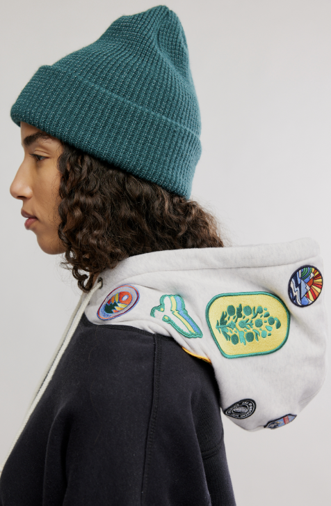 Person wearing a teal beanie and a jacket with colorful patches on a white background