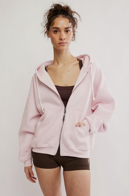 Woman wearing pink zip up sweatshirt with brown tank and shorts on plain background