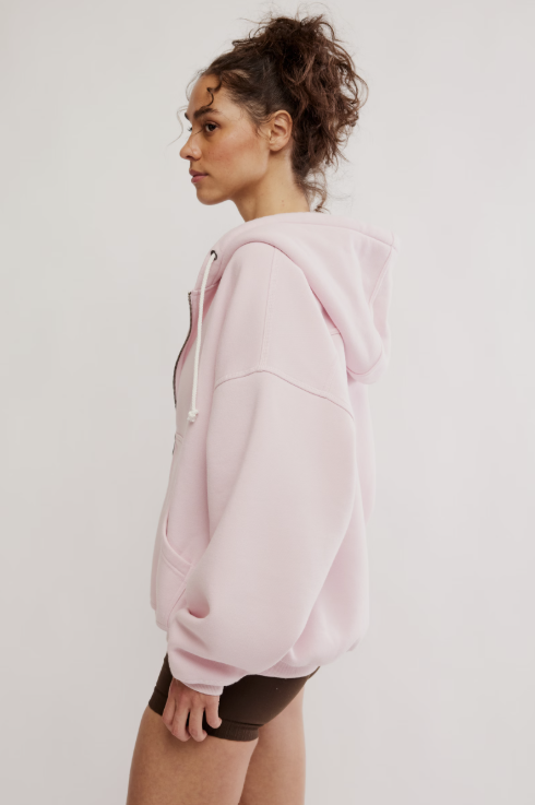 A woman in profile wearing a light pink oversized pullover hoodie with white drawstrings, paired with dark brown shorts, against a plain light gray studio background.