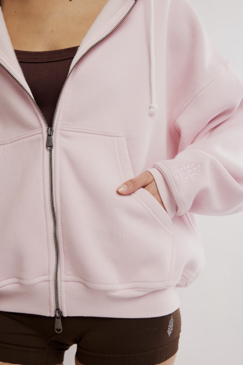 A close-up of a person wearing a light pink zip-up hoodie with white drawstrings, a dark brown ribbed tank top, and dark brown shorts against a plain light background.